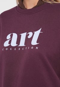 Felpa bordeaux con una stampa grafica bianca con la scritta "art COLLECTION." Ha un collo rotondo e una consistenza morbida.