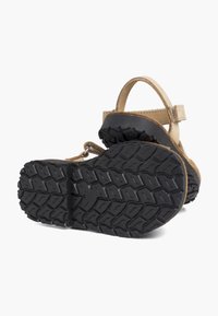 Vaaleanruskea nahkasandaali nilkkahihnalla, kulmassa, jotta paksu musta kumipohja, jossa on syvä kulutuspinta, näkyy.