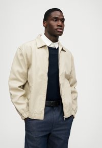 Bežs ar rāvējslēdzēju aizdare saspringts jaku ar elastīgām piedurknēm, vilkts pār tumšu džemperi un baltu kreklu ar apkakli, papildināts ar tumšiem džinsu biksēm un melnu jostu.