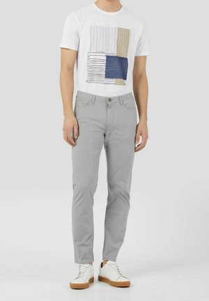 Mann trägt weißes Grafik-T-Shirt, hellgraue Slim-Fit-Hose und weiße Sneaker mit braunen Sohlen und steht vor einem schlichten Hintergrund.