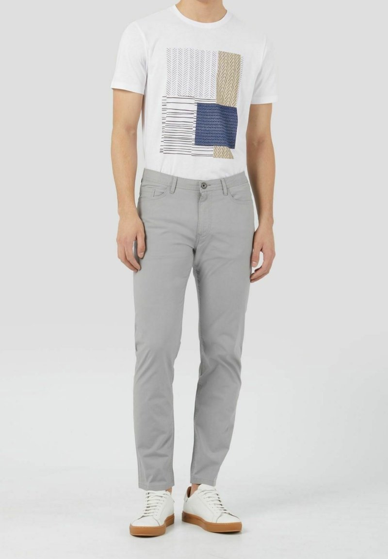 Homme portant un t-shirt blanc à motif graphique, un pantalon slim gris clair et des baskets blanches avec des semelles marron, debout devant un fond uni.