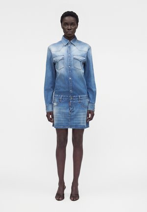 Μαύρη γυναίκα όρθια, φοράει ξεβαμμένο μπλε τζιν πουκάμισο με κουμπιά και ταιριαστό τζιν μίνι φούστα, καθώς και μαύρες ανοιχτές γόβες.