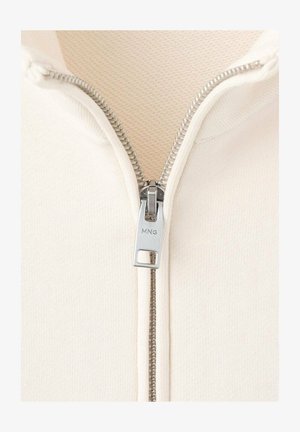 Close-up of silver zipper pull marked "MNG" on a cream-colored garment with visible zipper teeth and fabric texture.