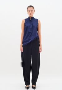 Bluză fără mâneci de culoare bleumarin, cu guler înalt, deschidere cu nasturi, combinată cu pantaloni largi negri și balerini negri cu vârf ascuțit, ținând o geantă mică.