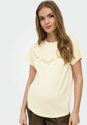 Jeune femme aux longs cheveux bruns portant un haut beige à manches courtes avec de la dentelle décorative et des pompons, posant devant un fond uni.