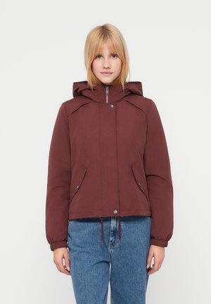 Ung kvinde iført en rødbrun hættetrøje med lynlås foran og trykknapper, kombineret med blå jeans, stående mod en hvid baggrund.