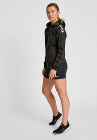 Zwarte hoodie met ritssluiting en capuchon, voorzien van witte chevron-accenten op de schouders. Gecombineerd met zwarte short en sportieve schoenen. Soepele textuur.