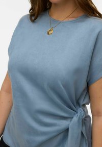 Haut en tissu doux bleu avec un détail noué sur le côté. Manches courtes et encolure ronde. Texture lisse, léger éclat.