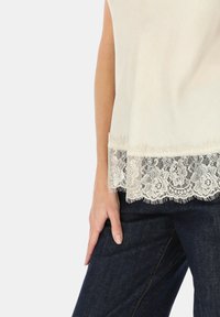 Bluzka w kolorze jasnej kości słoniowej z teksturowanej tkaniny, z koronkowym dołem i ozdobnymi frędzlami. Zestawiona z ciemnymi spodniami dżinsowymi.