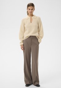 Sieviete valkā krēmkrāsas adītu džemperi ar atslēgas caurumu apakšā un plašu apkakli, kas apvienots ar augsto jostasvietu brūniem plaša griezuma biksēm un melniem apaviem.