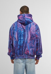 Uomo che indossa un hoodie multicolore con un motivo viola, blu e rosa, con il cappuccio alzato, abbinato a jeans di denim blu chiaro.