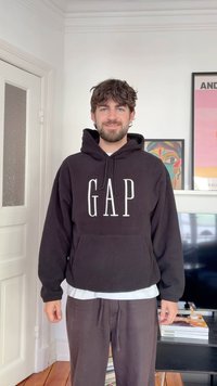 Young man with dark hair and beard wearing a black GAP hoodie and gray pants, standing indoors near a TV and framed artwork.
