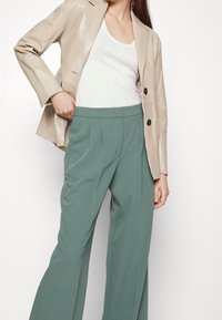 Blazer en cuir beige sur un débardeur blanc, associé à un pantalon large taille haute vert avec des plis et une texture lisse.