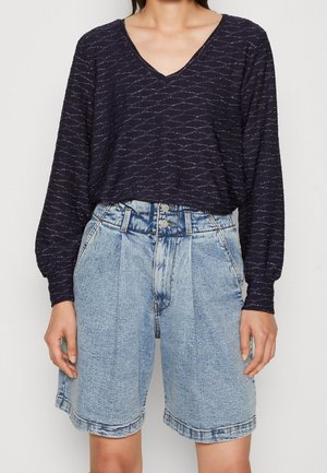 Vrouw die een marineblauw shirt met lange mouwen en V-hals draagt, met zilveren draadjes, en lichtblauwe denim shorts met hoge taille en voorplooien.
