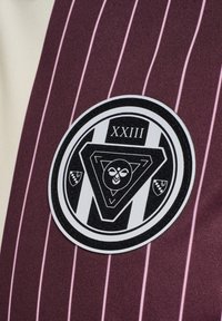 Bordowy materiał z pionowymi różowymi paskami ozdobiony okrągłą czarno-białą naszywką z geometrycznymi wzorami i napisem "XXIII".