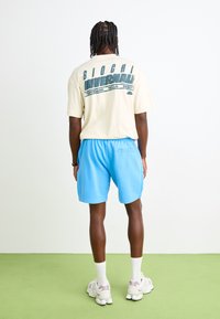 Tricou bej deschis cu text grafic turcoaz, asortat cu pantaloni scurți albaștri. Modelul poartă șosete albe și adidași gri deschis cu accente roz.