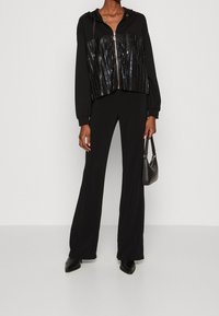 Felpa nera con zip e design lucido a costine sul petto, abbinata a pantaloni neri a gamba larga e scarpe nere con punta, mentre tiene una borsa piccola.