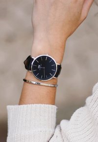 Black analog watch with a metal case, white hour markers, and a black leather strap, worn alongside a thin silver bangle on a light sweater.