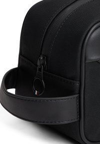 Black textured fabric bag with a leather strap, zip closure, and circular hardware accent. Compact rectangular design, minimalistic style.