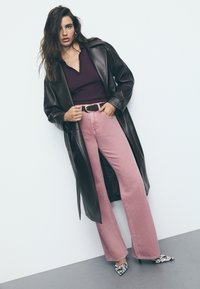 Smeđi kožni dugi kaput preko tamnoljubičastog topa, uparen sa ružičastim širokim trapericama i cipelama sa visokim potpeticama s uzorkom.