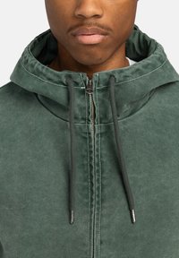 Chaqueta verde con cremallera y capucha, que cuenta con cordones ajustables, costuras blancas contrastantes y un acabado texturizado.