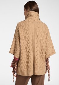 Poncho beige in maglia a treccia con colletto alto, orlo a coste e maniche ampie, che presenta un motivo testurizzato e una silhouette rilassata.