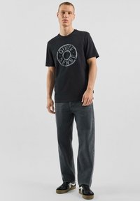 Czarny bawełniany t-shirt z okrągłym, szarym nadrukiem z napisem "QUITE SENSE"; w połączeniu z szarymi dżinsami i czarnymi sneakersami.