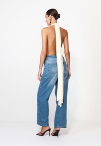 Zadný pohľad na osobu v krémovom topi s otvoreným chrbtom a dlhým volánom, v kombinácii s vysoko pásovými voľnými modrými džínsami a čiernymi podpätkami.