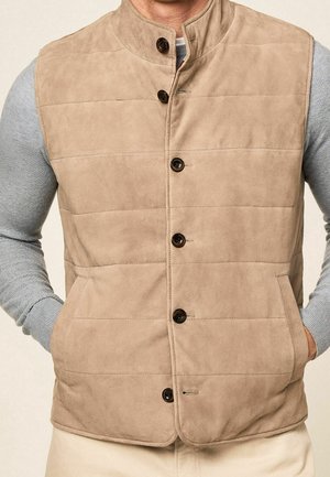 Beżowa pikowana kamizelka na guziki, noszona na jasnoszarym swetrze z długim rękawem, ręce w kieszeniach kamizelki.