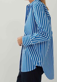 Modrá a bílá pruhovaná košile s dlouhými rukávy, zapínáním na knoflíky, špičatým límcem a zakřiveným lemem, vyrobená z lehkého materiálu.