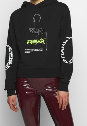 Hoodie noir avec texte blanc et jaune néon ainsi qu'un graphisme de casque audio, porté avec un pantalon en vinyle bordeaux brillant sur une personne debout.