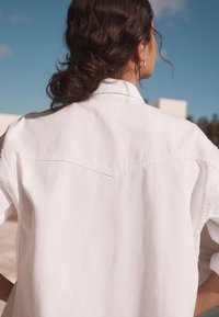 Camicia bianca oversize con bottoni, maniche corte, collo a punta e dettagli cuciti lungo l'imperdibile posteriore.