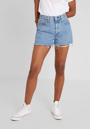 Person iført lyseblå slidte denimshorts, hvide høje sneakers og en hvid skjorte mod en ensfarvet grå baggrund.