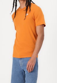 Oranžové bavlněné tričko s krátkým rukávem, kulatým výstřihem a malou bílou nášivkou na hrudi. V kombinaci s modrými džíny.