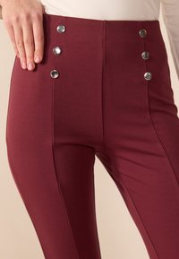 Pantalons bordeaux taille haute avec une texture lisse, présentant cinq boutons métalliques argentés sur le côté et des détails de couture le long des jambes.