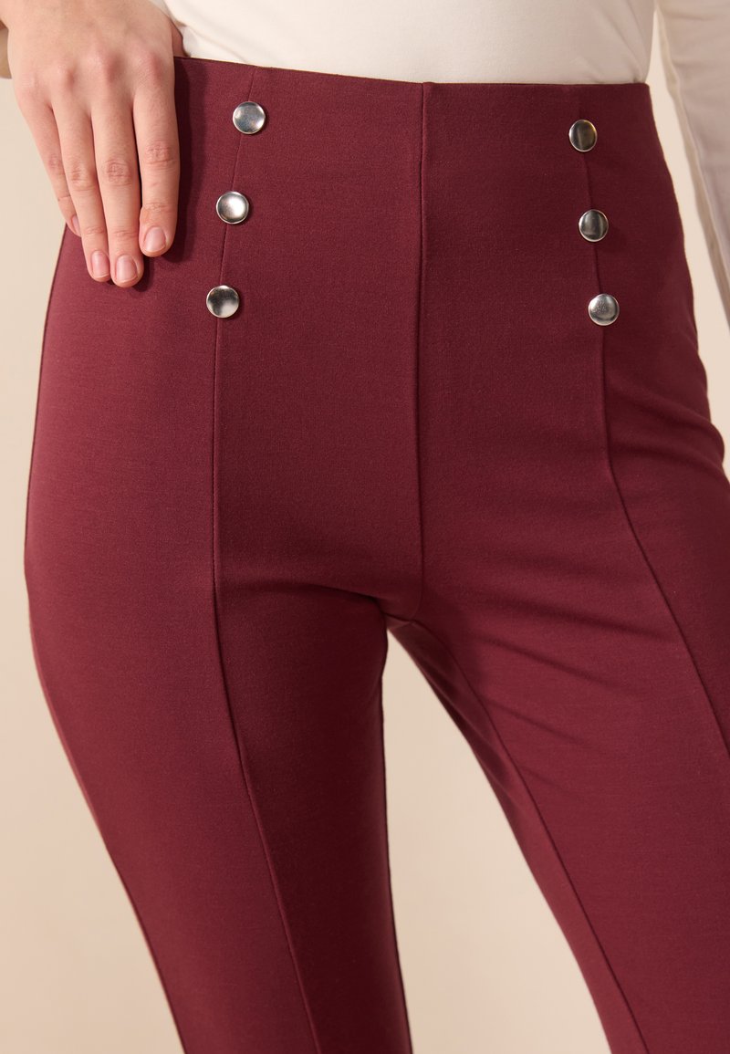 Pantaloni a vita alta bordeaux con una texture liscia, caratterizzati da cinque bottoni metallici argentati sul lato e dettagli di cucitura lungo le gambe.