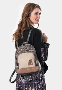 Mujer con cabello largo y castaño sonríe, vestida con una chaqueta negra y una falda de estampado, llevando una mochila Anekke en tonos marrón y beige sobre su hombro.