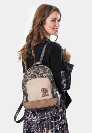 Mujer con cabello largo y castaño sonríe, vestida con una chaqueta negra y una falda de estampado, llevando una mochila Anekke en tonos marrón y beige sobre su hombro.