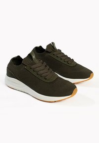 Dunkelgrüne Strick-Sportschuhe mit einer weißen Zwischensohle und einer Sohle aus Naturkautschuk. Merkmale sind runde Schnürsenkel und ein strukturiertes Obermaterial.