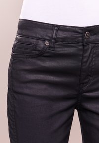 Jean en denim noir avec une texture lisse, présentant un design classique à cinq poches, des éléments en métal argenté et des détails de couture subtils.