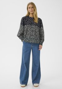Woman with long blonde hair wearing a dark floral buttoned blouse, wide-leg blue jeans, and white sneakers standing against a white background.