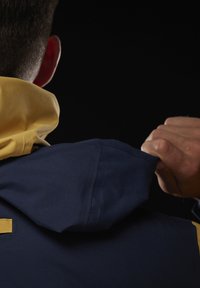 Námořnická modrá bunda s kapucí a žlutým límcem, ukazující texturu látky a detaily šití. Ruka držící límec pro úpravu.