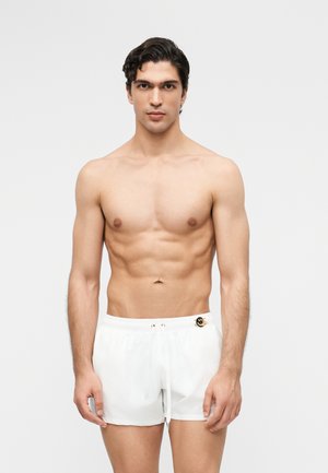 Mladý muž s krátkymi tmavými vlasmi, oblečený v bielych plavkách s čiernym emblémom usmievajúcej sa tváre, stojaci na obyčajnom svetlom pozadí.