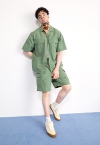 Green button-up shirt with two front pockets, matching shorts, and beige sneakers; set against a white background and blue floor.