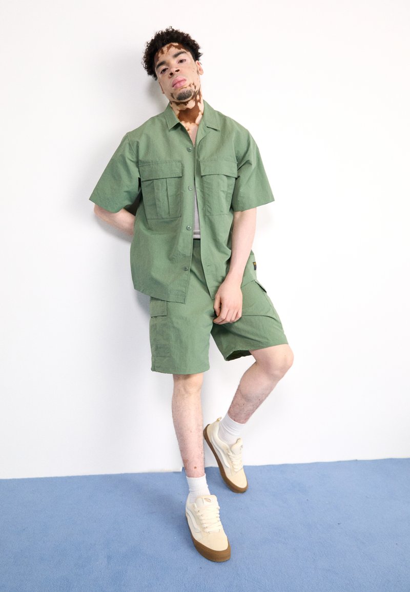 Camisa verde de botones con dos bolsillos delanteros, pantalones cortos a juego y zapatillas beige; presentado sobre un fondo blanco y un suelo azul.