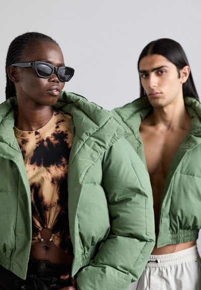 Dwie modelki noszą oversize'owe, puchowe zielone kurtki. Kurtka jednej modelki ma wysoki kołnierz; obie prezentują lekkie, teksturowane materiały.