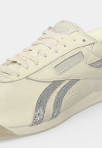 Baskets Reebok blanc cassé avec lacets blancs et bandes argentées texturées sur le côté, affichant le nom de la marque sur la languette et le panneau extérieur.