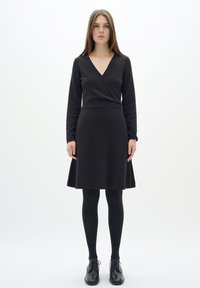 Abito nero avvolgente con scollo a V, maniche lunghe e gonna svasata; abbinato a collant neri e scarpe. Tessuto dalla consistenza morbida.