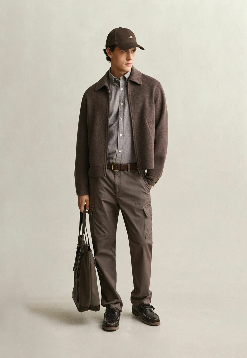 Hombre con chaqueta marrón, camisa gris abotonada, pantalones cargo marrones, zapatos oscuros y gorra, sosteniendo una bolsa tote marrón, de pie sobre un fondo claro.