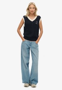 Maglione senza maniche blu navy con scollo a V color avorio e texture a trecce; abbinato a jeans denim chiari larghi e sneakers grigie.